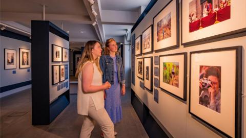 Two women with blonde hair in an art gallery looking at photographs of the British royal family on a wall.