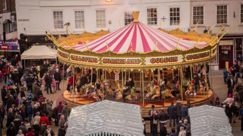 People gathered around a carousel, with market stands and shop fronts in the background. 