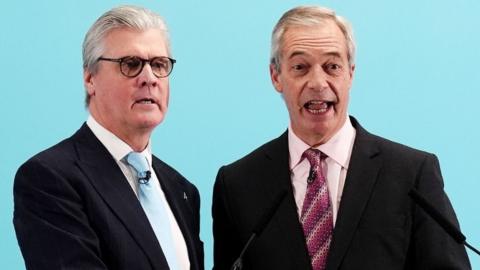 Malcolm Offord, who has grey hair and glasses, next to a smiling Nigel Farage, who has thinning grey hair. Both are wearing dark suits, and standing in front of a pale blue background. 
