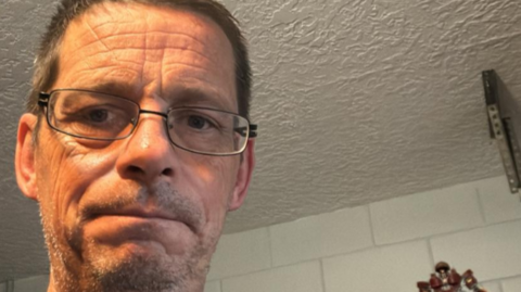 A selfie of a man wearing glasses who has short hair and stubble. He is looking towards the camera with a worried expression. He is standing in front of a white tiled wall