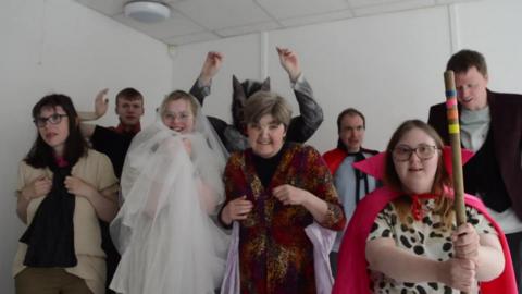 A group of people dressed up in spooky costumes, including a ghost and a werewolf. They are all smiling, some with their hands in the air.