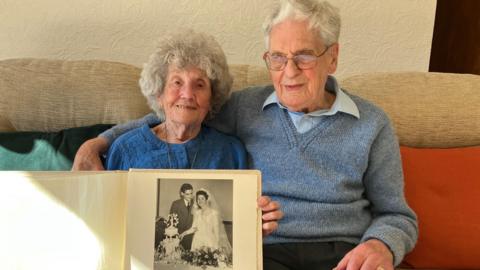 An elderly couple sit on a cream sofa together while the woman in a blue top holds up a photo album showing a black and white photo of them when they were younger cutting their wedding cake. The man is sitting in a grey jumper with his arm around the woman. 