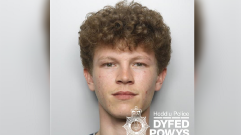 A mugshot of a teenager with curly brown hair and hazel eyes. Against a white wall.