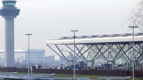 Stansted Airport, showing the tall control tower and white roof of the terminal building. Cars are arriving at the drop-off at the terminal, with people entering the building.