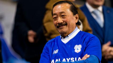 Vincent Tan in a blue Cardiff City shirt smiles with arms folded in the stands at a Cardiff City games