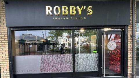 The outside of the restaurant with a black front and gold sign saying "Robby's Indian Dining"