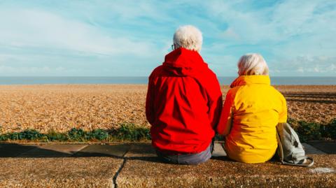 Two grey-haired people sitting with their backs towards the camera. They are sitting on a low wall with a pebbly beach and the sea in front of them. One is wearing a bright red rain coat and the other a bright yellow.