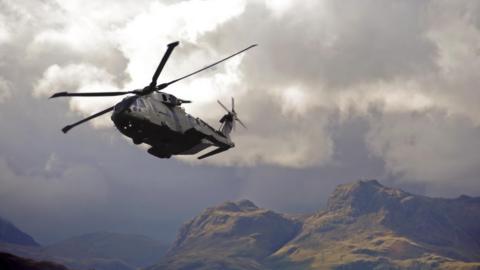 An RAF helicopter above a mountain range - it flies low to the cliff face against a dramatic grey cloud.