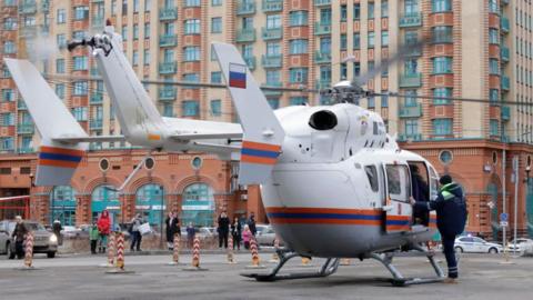 A man climbs into a white medical helicopter. A red-brick building towers in the background