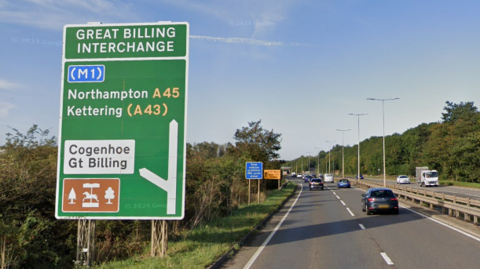 The A45 at the Great Billing Interchange junction. There is a large sign showing motorists the left turn for the junction. The sign has directions to Northampton, Kettering, Cogenhoe and Great Billing. To the right of the sign is the dual-carriageway road, which has many cars on it.