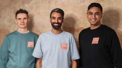 The trio are wearing light coloured T-shirts or tops with their branding on - an orange square with Padel Warehouse 52. They are smiling
