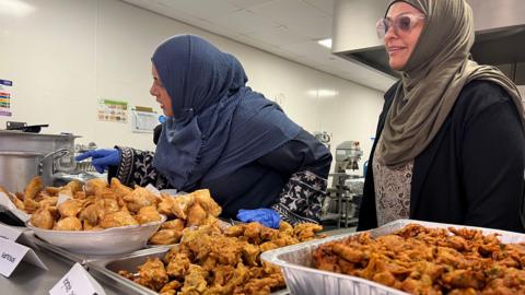 Two women standing over large silver trays of dark gold, crisp-looking fried food in a kitchen.