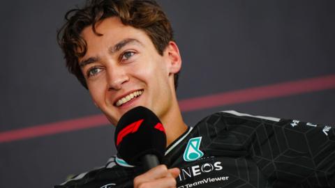 A smiling George Russell speaking into an F1-branded microphone during a news conference at Bahrain testing