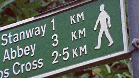 Picture of a green signpost, with a logo of a figure walking on the right hands side.  The text reads: 'Stanway 1 KM', (the word 'Hailes' is obscured) partially visible '..Abbey 3 KM' visible and (the word 'Stumps' obscured) visible 'Cross 2.5 KM'