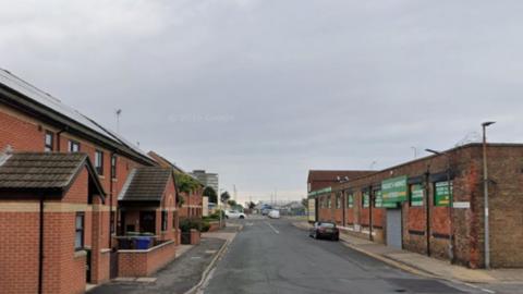 Section of Weelsby Street in Grimsby. There is a single storey factory building on the right and red brick houses on the left