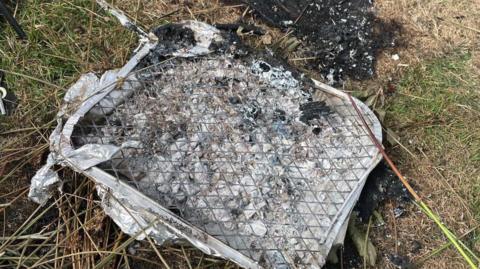 The charred remains of a disposable barbecue left on Dartmoor with severe black burn marks on the parched grass beside it.