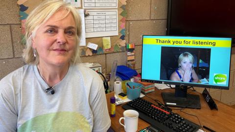 A woman with blonde hair sat at a desk next to a computer which has an image of her daughter on and the words  "thanks for listening" above the photo. On the desk are a coffee cup and some stationary, including pens, paper and a ruler. 