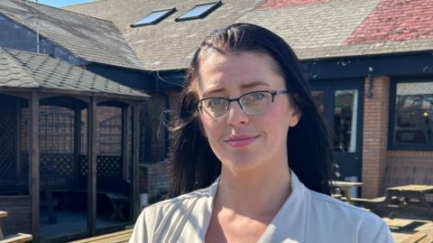 Chanel Walsh standing near a pub garden. There are benches behind her and there is a St. Georges flag on the roof. Behind her there is a shelter.