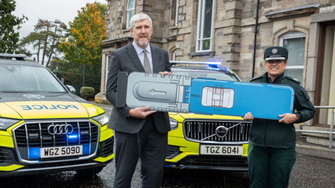 John O'Dowd in a black suit and a grey shirt holds a large drug testing kit alongside a female police officer, in uniform, in front of two police cars