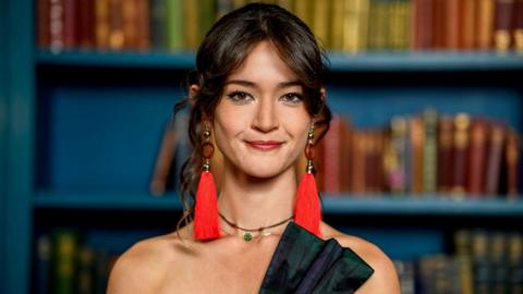 Woman with long dark hair which is pinned up behind her head with a long fringe at the front brushed away to each side, she is smiling and wearing long red tassel style earrings and two delicate necklaces, one of which has a green gemstone. The off the shoulder outfit is in navy and dark green tartan styled like a bow on the right side. She is standing in front of a blue bookcase which has books on the shelves.