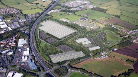 An aerial view of the proposed industrial site which is a large flat building to the right of the M6. There are other industrial unit buildings on the land, which is surrounded by fields and trees. On the left of the M6 is a more built up area. 
