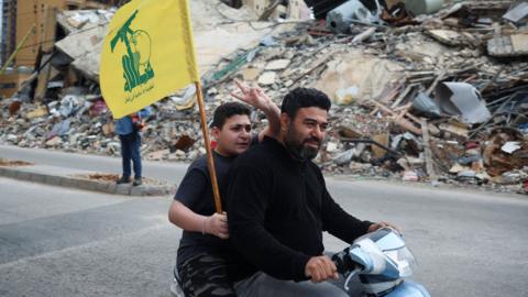 Two people ride on a moped, while one holds a Hezbollah flag and holds two fingers in the air, on a street in Beirut in front of a huge pile of rubble where a building once stood, taken on Friday.