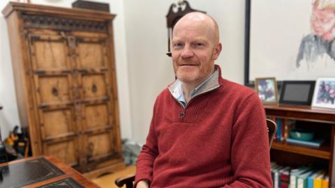 A  man in his 50s with red hair and  beard, wearing a red jumper. He is sitting at a desk. He wears a red jumper and  a blue shirt. 