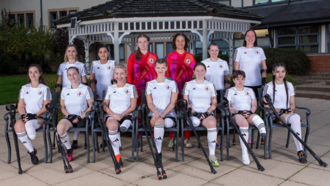 Seven women from the amputee football team sit on chairs, while six more members of the team stand beside them. They all wear an England football kit and smile at the camera. The kits are white, apart from two players who are goal keepers, who wear a red and pink kit. They are all outside in front of a pagoda.