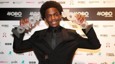 DC3 holding up the best newcomer and best gospel act awards, he has one in each hand. He is wearing a black suit, with black shirt and black tie. He is standing in front of a wall with Mobo Awards and other writing and logos on it. 