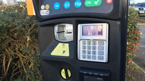 A black parking machine, with numbers and other functions on keys and a slot for cards to be inserted, with bushes behind it