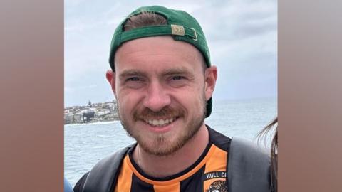 Jack Gorbert pictured wearing a Hull City shirt and a green baseball cap. There is an expanse of water in the background.