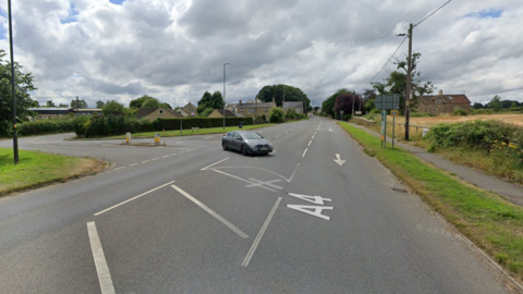 A lone car uses the junction of the A4 and Bradford Road in Corsham. Trees, fields and street furniture appears in the background.