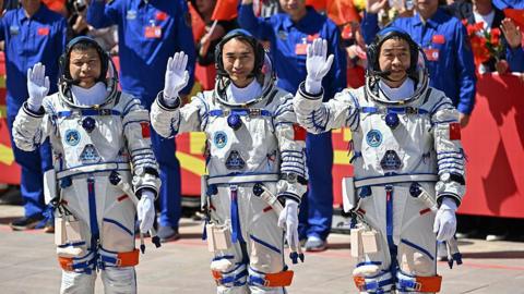 Wang Jie, Chen Zhongrui, and Chen Dong stand in space suits waving at a crowd of people.