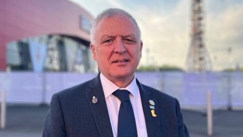 A man with short grey hair wears a navy suit, white shirt and navy tie. He has three badges on his left lapel and one on his right. Over his right shoulder is a large red building which is out of focus. It is day time, the sky is blue, partially cloudy. 