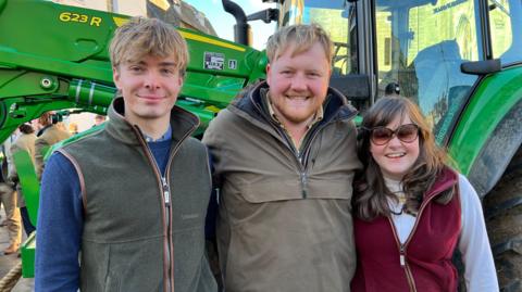 Two agriculture students - a young man and a young woman - stand either side of Clarkson's Farm star Kaleb Cooper as they stand in front of a green tractor in Cirencester town centre. They are all smiling.