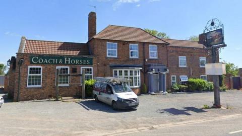 A two-storey pub sits at the side of a road.  The name Coach & Horses is written on the building