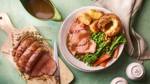 A cooked roast sits on a chopping board along with a knife which has clearly sliced some cuts already. Beside the roast is a plate filled with roast meat, potatoes, greens and Yorkshire pudding.