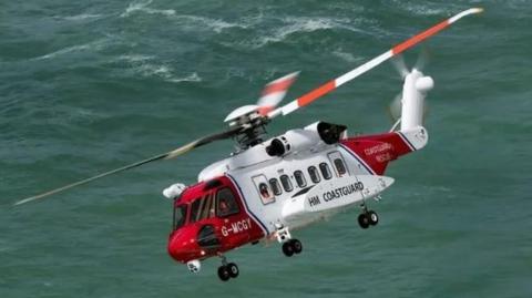 A white and red HM Coastguard helicopter flying over the sea