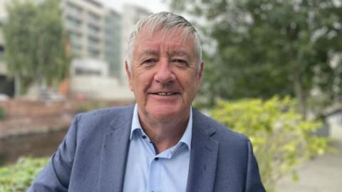 A man with grey hair and a blue suit with an open necked shirt in an outdoor area