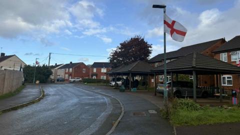 A St George's flag flies in the wind fixed to a lamppost on the inside edge of a curved pavement. Along the right side of the image there are orange bricked houses and cars parked on driveways. There is a bright blue sky blotted with dark grey clouds.