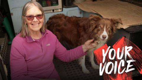 A woman in a pink fleece and wearing glasses sits in the back of a van with her border collie. 