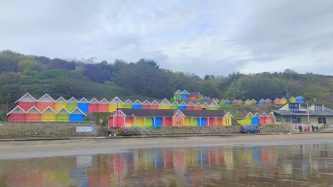 three tiers of colourful beach huts reflected in the damp sand, large trees behind them, under a cloudy sky