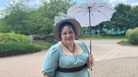 Sophie Andrews stands outside in her regency outfit and looks at the camera, smiling. She has dark brown curly hair, with two strands of ringlets framing her face and the rest of her hair in an up-do underneath a straw bonnet which has a pale blue ribbon rim and blue feathers sticking out the top. Sophie holds a parasol made of white lace. She is wearing a brown stone necklace with a turquoise blue dress, which has three quarter length sleeves and a brown ribbon sash around her waist. The background is blurred but shows trees and hedging surrounding a pale beige cobbled area of road.