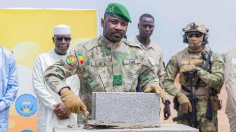 Gen Assimi Goïta, in military uniform, is standing in front of a brick at the construction site of the refinery in Senou, as armed soldiers in uniform and men in civilian clothes look on