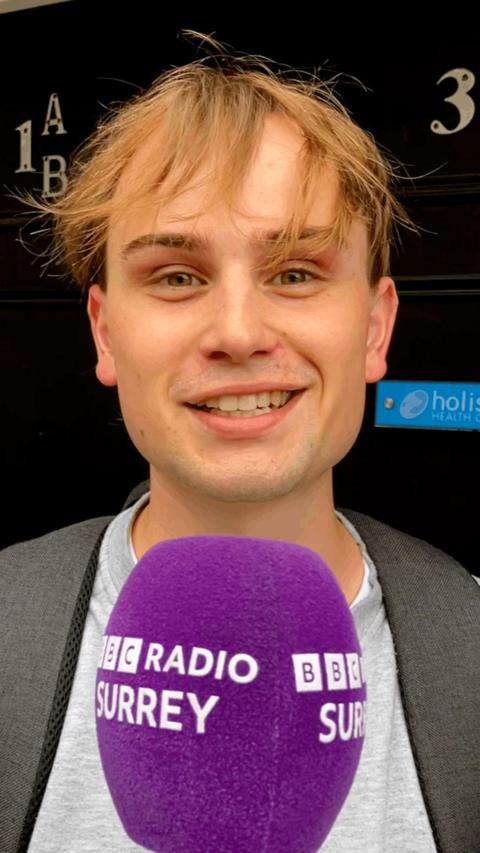 A young man with long, brown hair standing in front of a purple microphone.