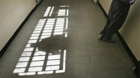 A corridor with sunlight streaming through a barred window, casting shadows on the tiled floor. A person stands against the right wall, only their lower body visible. Their shadow on the floor.