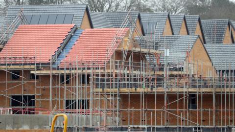 A set of houses being built with a variety of black and brown tiled roofs and lots of scaffolding poles around them.