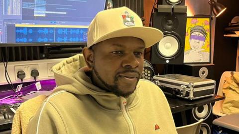 Xidus Pain in a recording studio. He is wearing a yellow hoodie with a yellow baseball cap and has a short neat black beard. Behind him is a mixing desk, speakers and a computer screen.