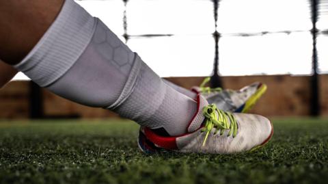 A close-up of someone's legs with white football socks and boots. They are sitting on a green artificial pitch.