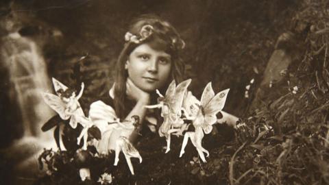 A black and white photo of a young girl with brown hair and a headband resting her hand on her neck and looking at the camera. In front of her are small dancing fairies. She is sitting outdoors with what appears to be a stream behind her.